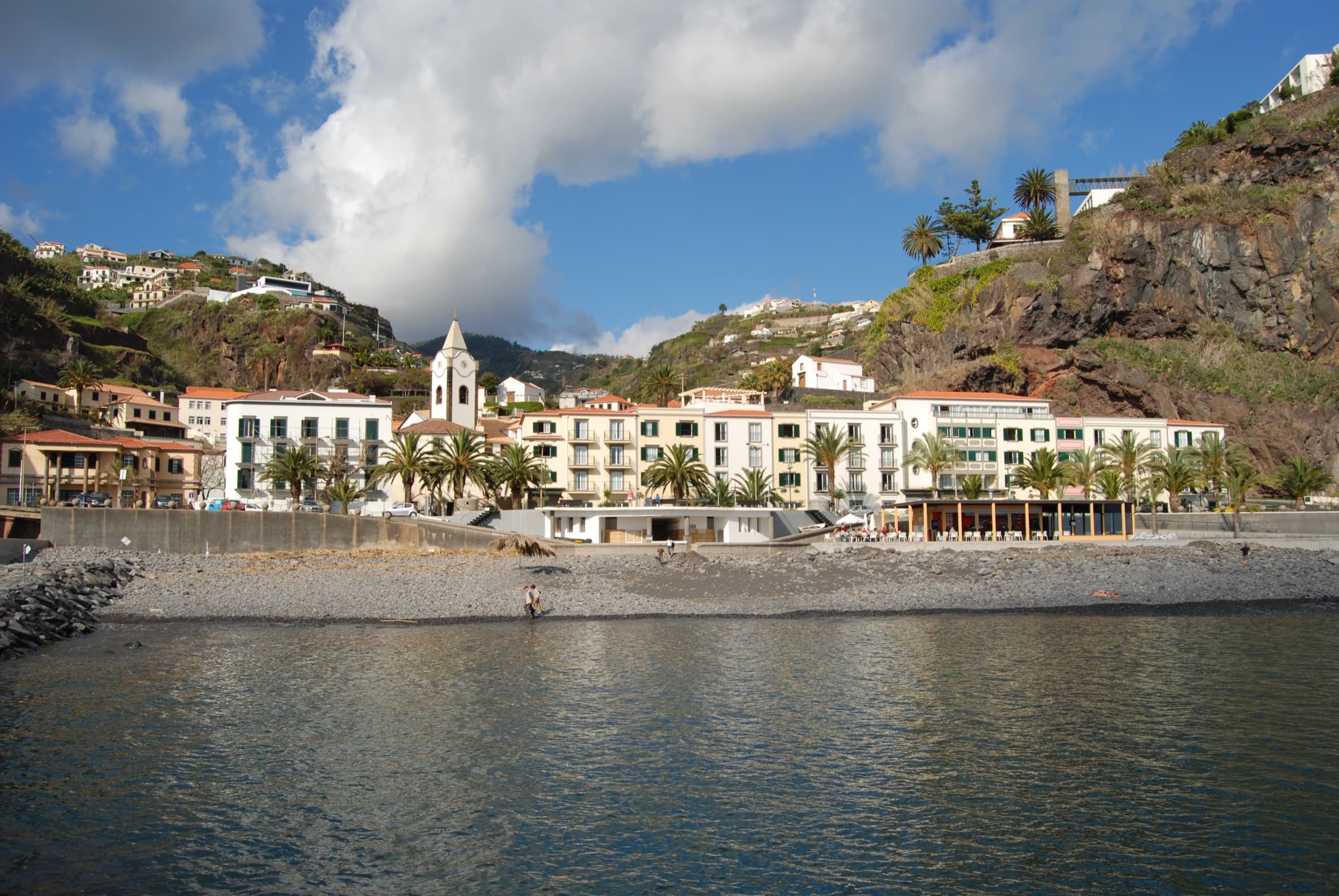 The white seafront buildings and church tower of Ponta do Sol sit above a pebble beach, with green cliffs behind.