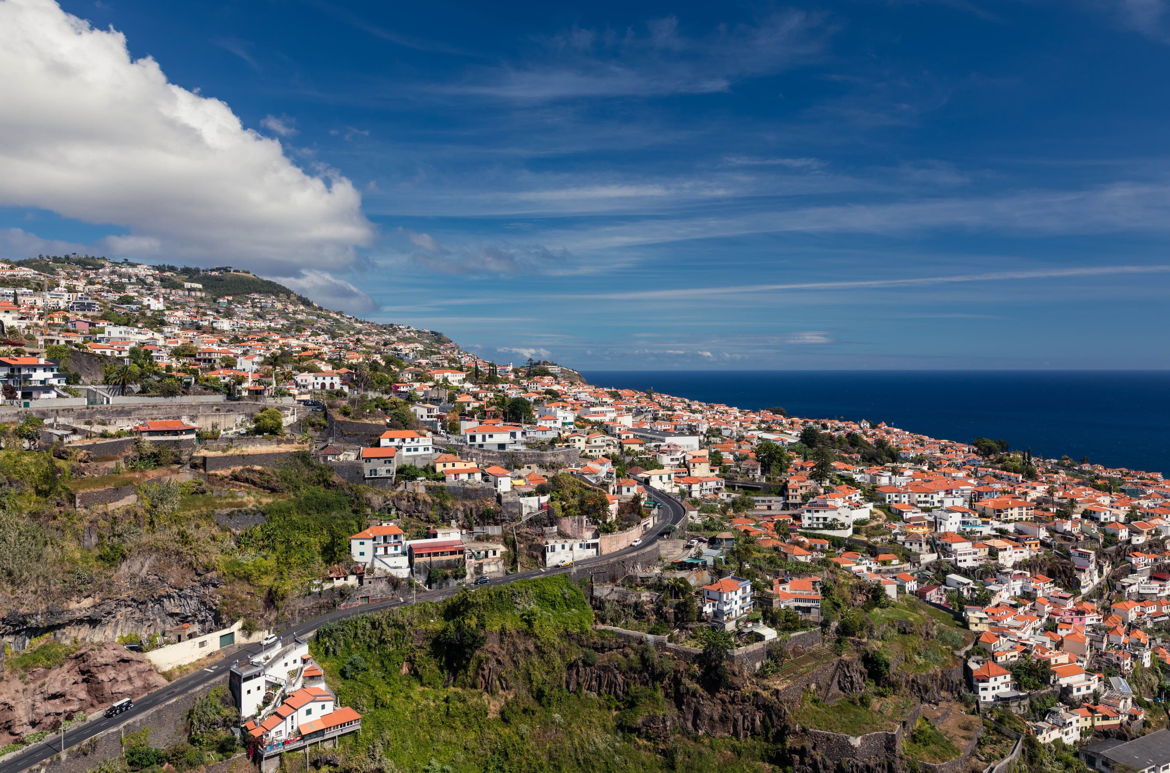 Funchal’s red-roofed houses climb the green hillside above the Atlantic under a bright blue sky.