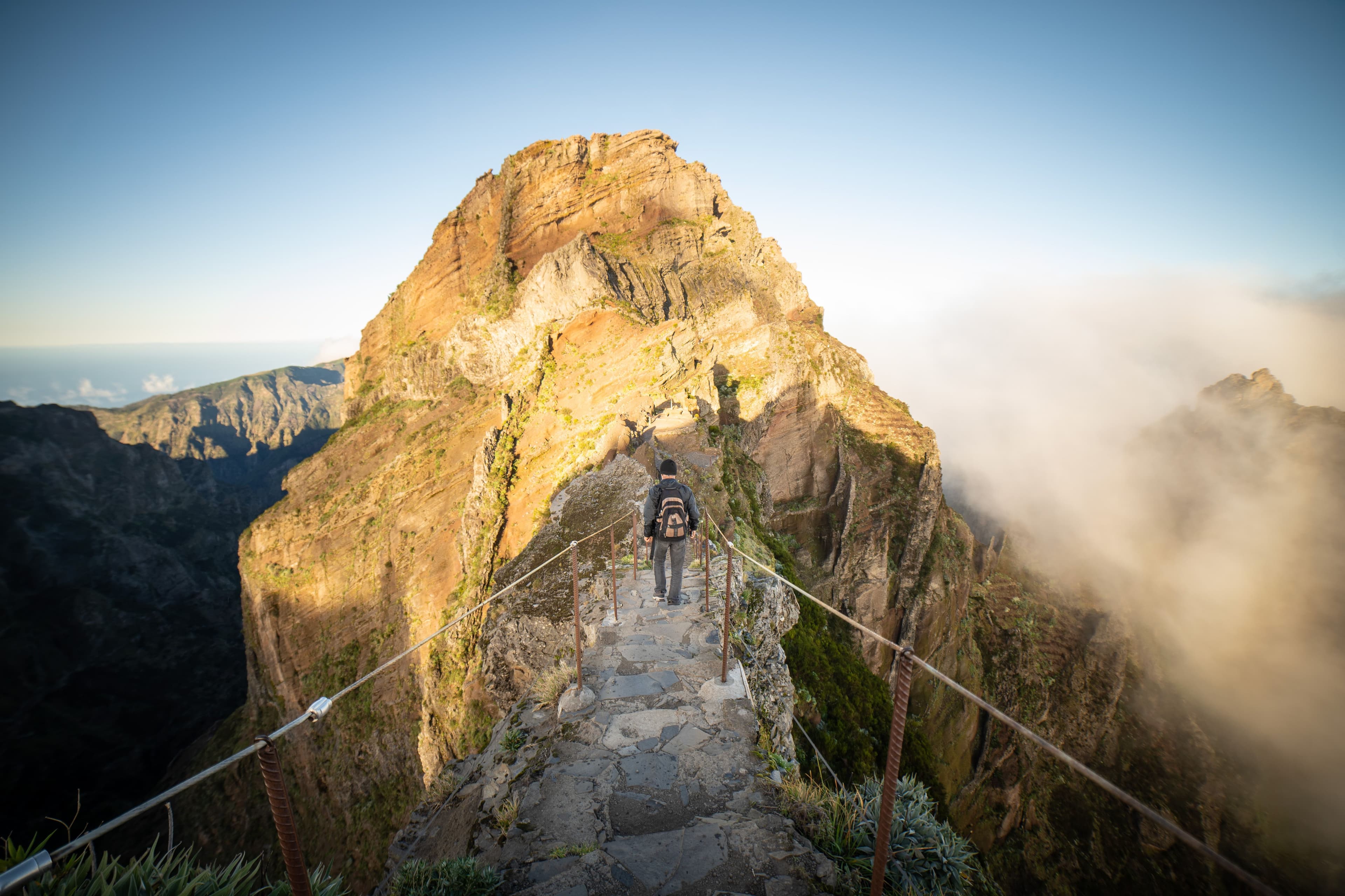 A hiker climbs a stone ridge path lit gold by sunrise as clouds swirl below, between Pico do Arieiro and Pico Ruivo.